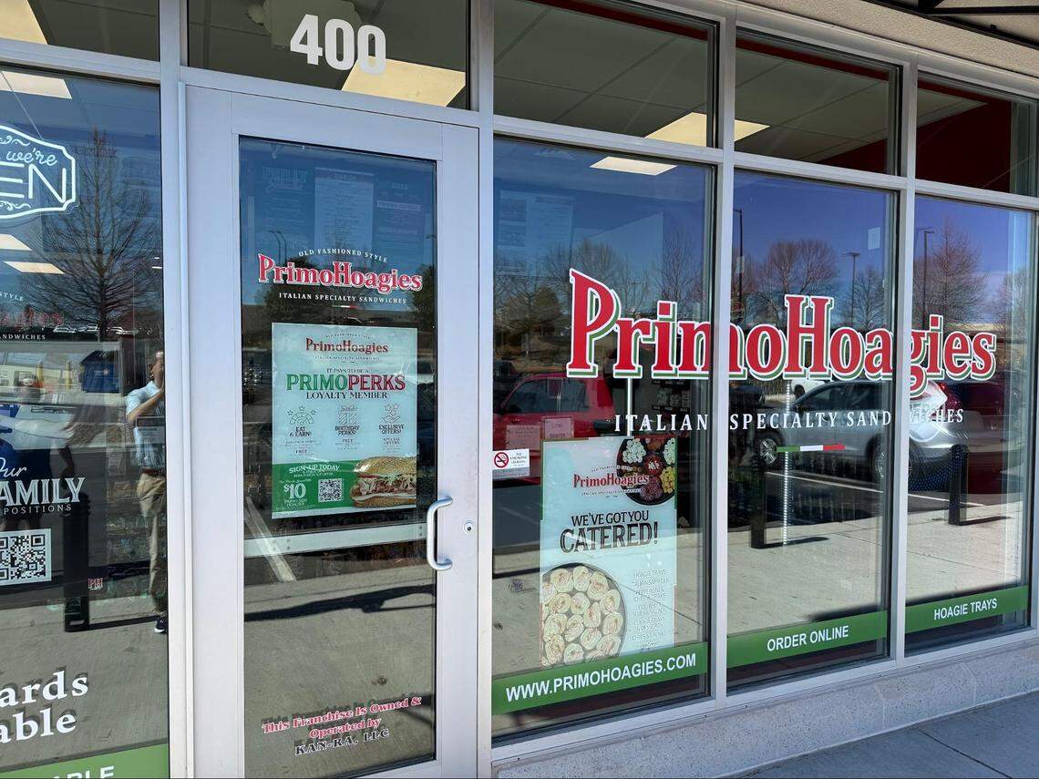 A close-up of the glass entrance door to a PrimoHoagies storefront. The door is adorned with the restaurant’s logo and several promotional posters, including one for “PrimoPerks” and another for catering. The reflection of the parking lot is visible in the glass.