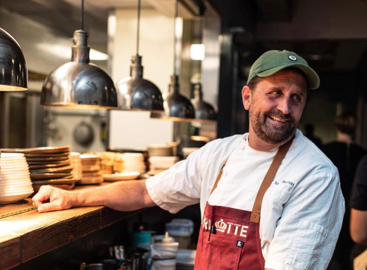 A portrait of a chef, smiling and bearded, in a backwards baseball cap and a red apron, resting an arm on the thick wooden counter of the kitchen pass, under a line of steel heat lamps, with stacks of dishes ready for service.