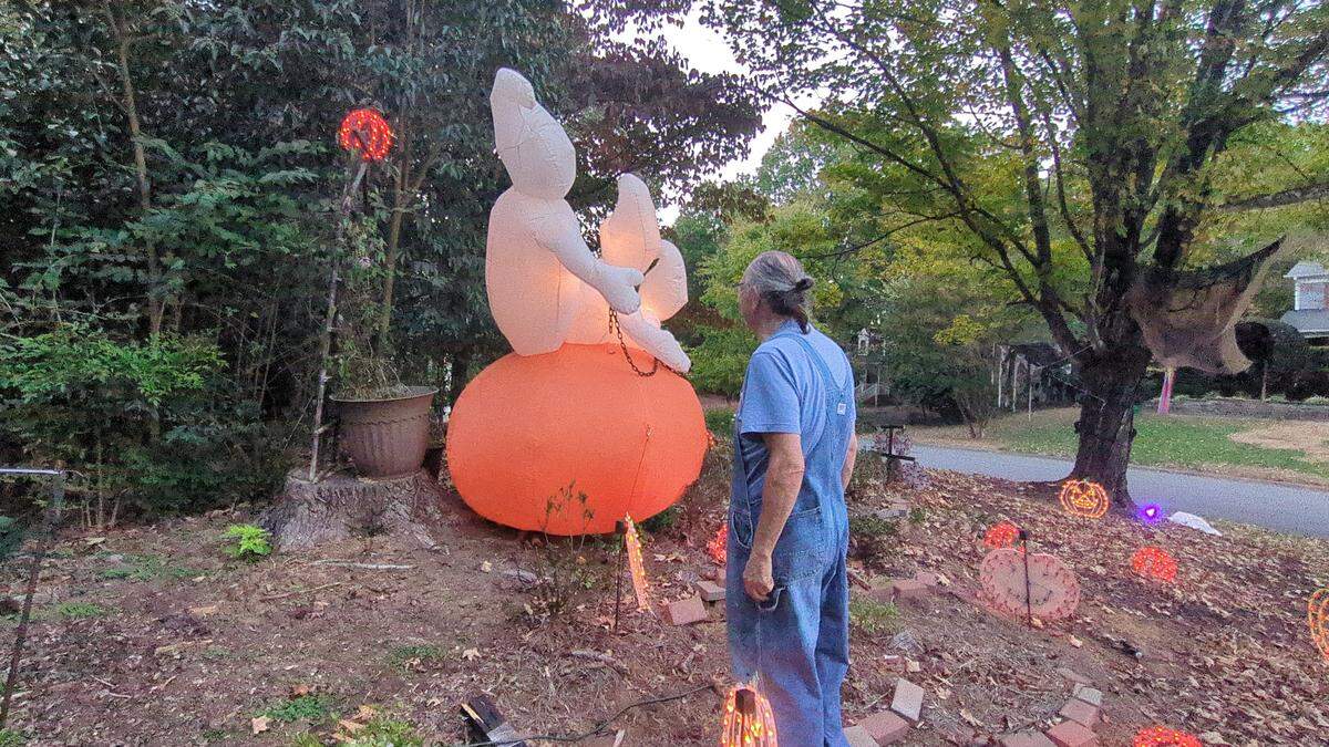 A wide shot of a man in denim overalls standing in a yard and looking at a large, illuminated, inflatable Halloween decoration. The decoration features a white ghost sitting on top of a large orange pumpkin. The yard, pictured at dusk, is also decorated with smaller, light-up pumpkins and is surrounded by trees.