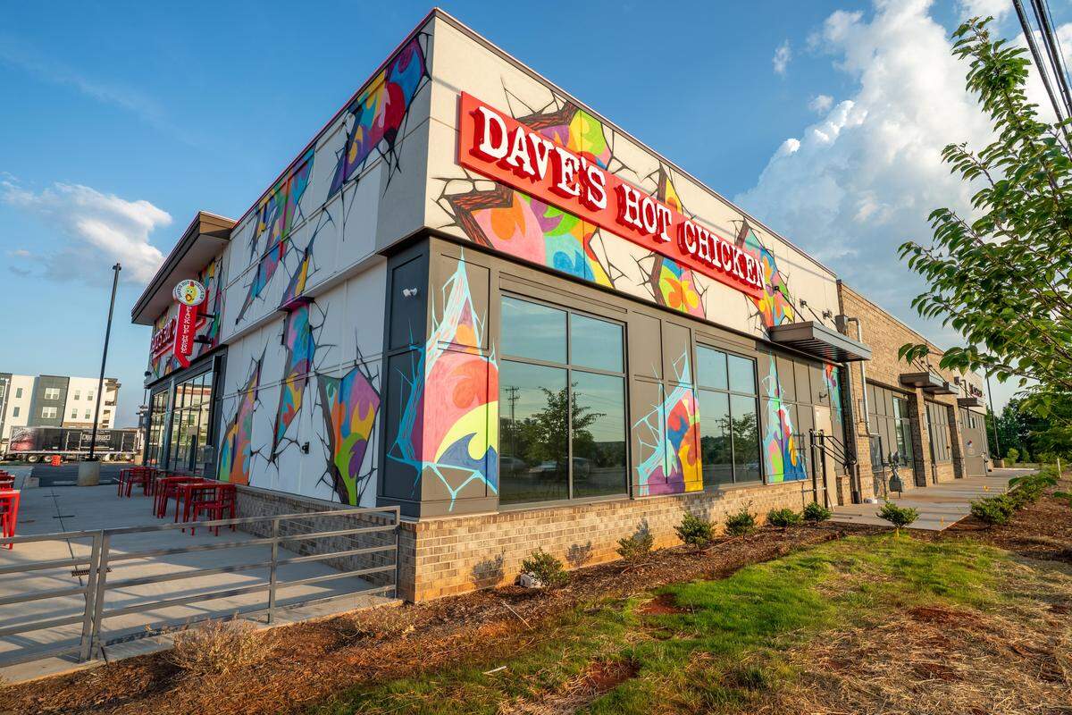 A low-angle, outdoor shot of a modern “Dave’s Hot Chicken” restaurant storefront under a bright, partly cloudy sky. The building’s exterior features a light beige and dark gray facade heavily adorned with colorful, graffiti-style murals that look like vibrant geometric patterns bursting through cracked stone. A prominent red channel-letter sign reading “DAVE’S HOT CHICKEN” is mounted on the front corner. Large glass windows line the side of the building, and a small outdoor patio with red tables and chairs is visible behind a metal railing on the left.