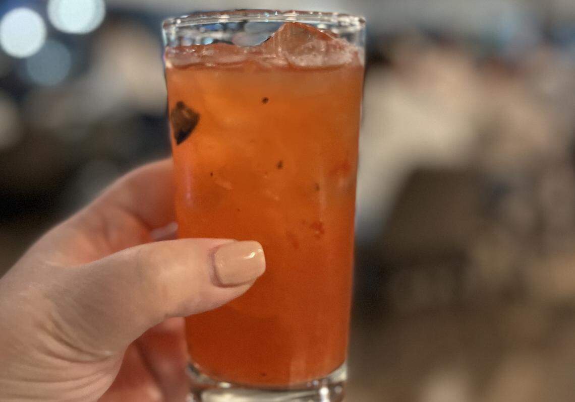 A close-up shot shows a hand with light-colored nail polish holding a tall, clear glass filled with a bright pink, icy cocktail. The background is completely blurred with soft bokeh lights.