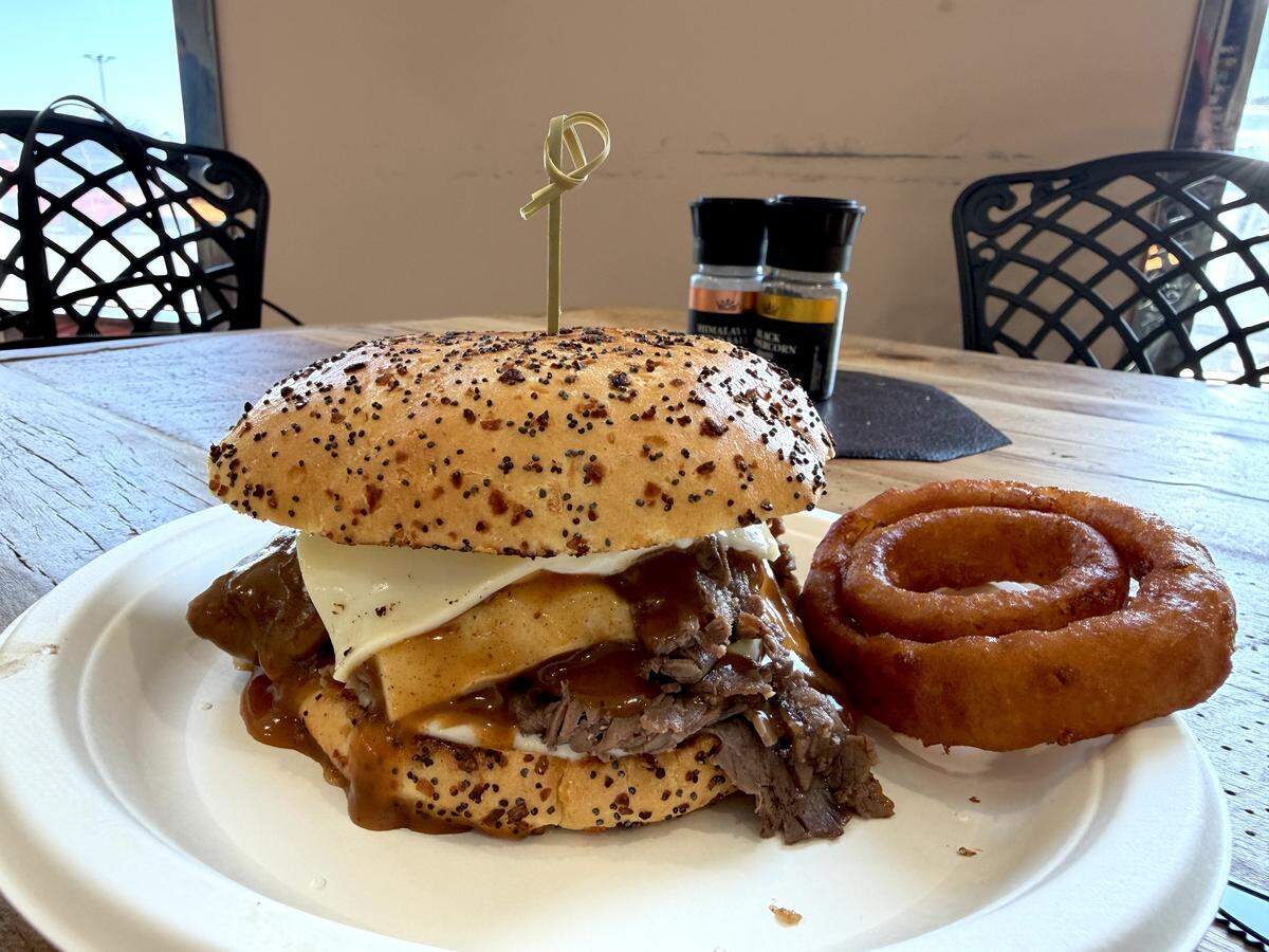 A gourmet roast beef sandwich on a toasted everything-seeded bun, dripping with brown gravy and topped with melted white cheese, served with a large, golden-brown battered onion ring on a white plate.