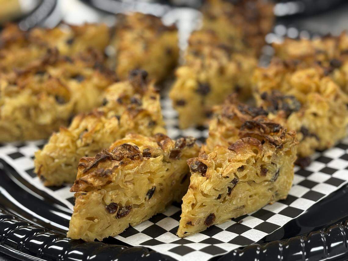 A detailed, close-up shot of a platter of kugel. The kugel is cut into square portions, showing a dense, baked texture with wide egg noodles and dark raisins, featuring a crispy, golden-browned top layer.