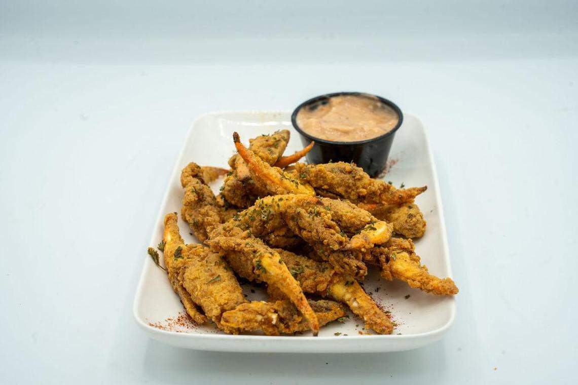 A mound of golden-brown, breaded and fried crab claws piled on a white rectangular plate. A small black ramekin of creamy dipping sauce sits in the corner, and the dish is lightly dusted with paprika and dried parsley.