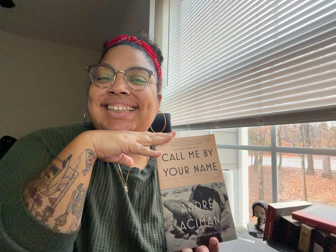 A smiling person with glasses, a red bandana, and visible tattoos on their arm is holding the book “Call Me By Your Name” by André Aciman, resting their chin on their hand near a window with horizontal blinds.
