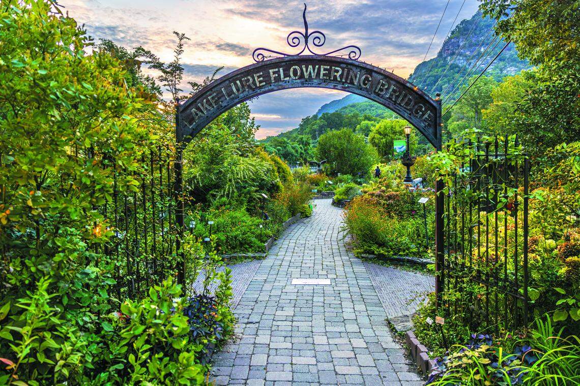 A wrought-iron archway reading “Lake Lure Flowering Bridge” stands over a stone paver path, flanked by lush green gardens. In the background, a mountain rises against a cloudy sunset sky.