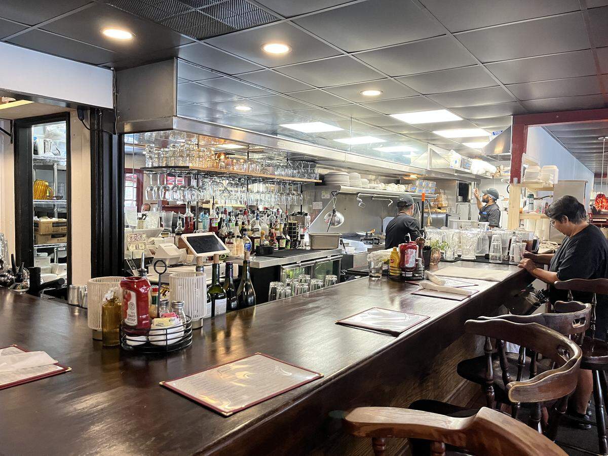 Looking along the length of a polished dark wood bar in a diner-style restaurant. Menus are laid out on the counter in front of empty barstools. At the far end, a patron is seated. The area behind the bar is busy, with an overhead rack full of clean wine glasses, numerous liquor bottles, and staff working in the visible kitchen area.
