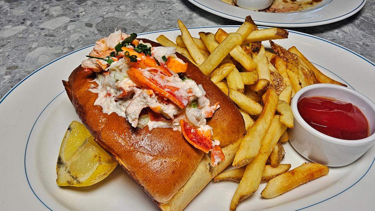 A slightly elevated close-up shows a large white oval plate with a blue rim holding a generous lobster roll and a side of golden-brown french fries. The lobster roll is made with a toasted, buttered split-top bun overflowing with chunks of bright orange and white lobster meat mixed in a creamy dressing, garnished with fresh green chives. A lemon wedge sits next to the lobster roll. The french fries are piled neatly next to a small white ramekin filled with red ketchup. The plate rests on a gray mottled countertop, and another blurry plate is visible in the upper right background.