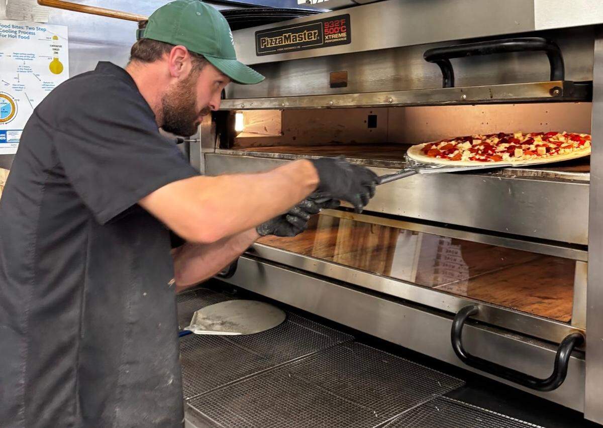 A medium shot shows a pizza chef wearing a black shirt, green cap and black gloves from the side, using a metal peel to slide an uncooked pizza with sauce and toppings into the top level of a large, multi-deck, stainless steel “PizzaMaster” oven.