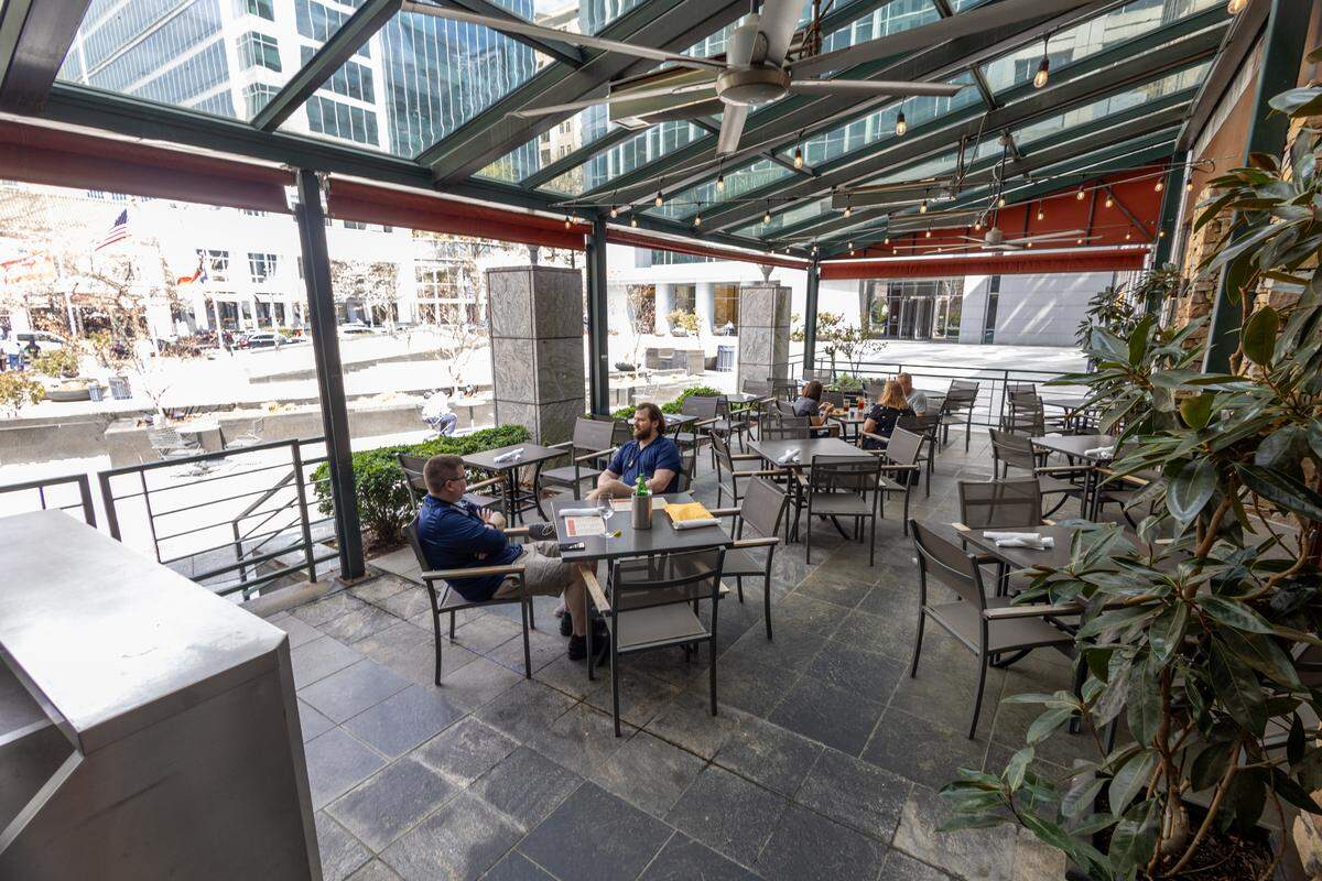 A bright, glass-enclosed patio with a view of a city plaza. Several patrons are seated at dark metal tables. The space features a high glass ceiling with industrial fans, string lights, and large potted greenery.