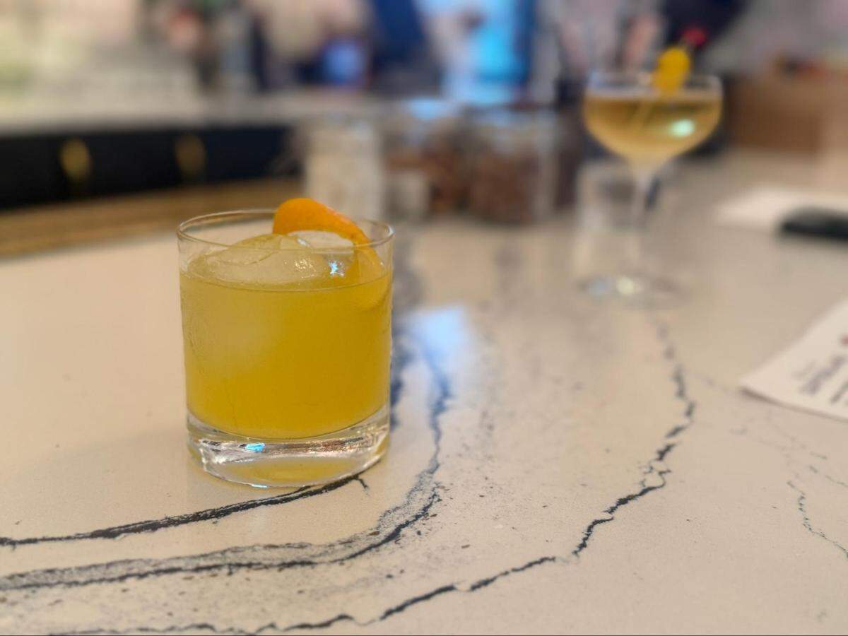 A close-up shot of a yellow-orange cocktail in a rocks glass, garnished with an orange peel, sitting on a white and gray marbled countertop. The drink is in sharp focus in the foreground, while the background is blurred, showing a bar setting with another cocktail in a coupe glass further down the counter.