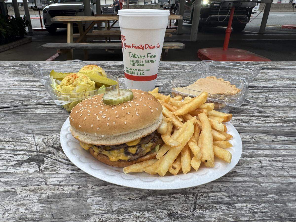 A full meal spread from a drive-in restaurant laid out on a rustic wooden picnic table. In the center, a white plate holds a double cheeseburger with a sesame seed bun topped with a pickle slice, alongside a large pile of golden french fries. Behind the plate is a tall white styrofoam cup reading “Your Family Drive-In.” Flanking the plate are two clear plastic takeout containers: one with potato salad and a pickle spear on the left, and one with a slice of pie on the right.