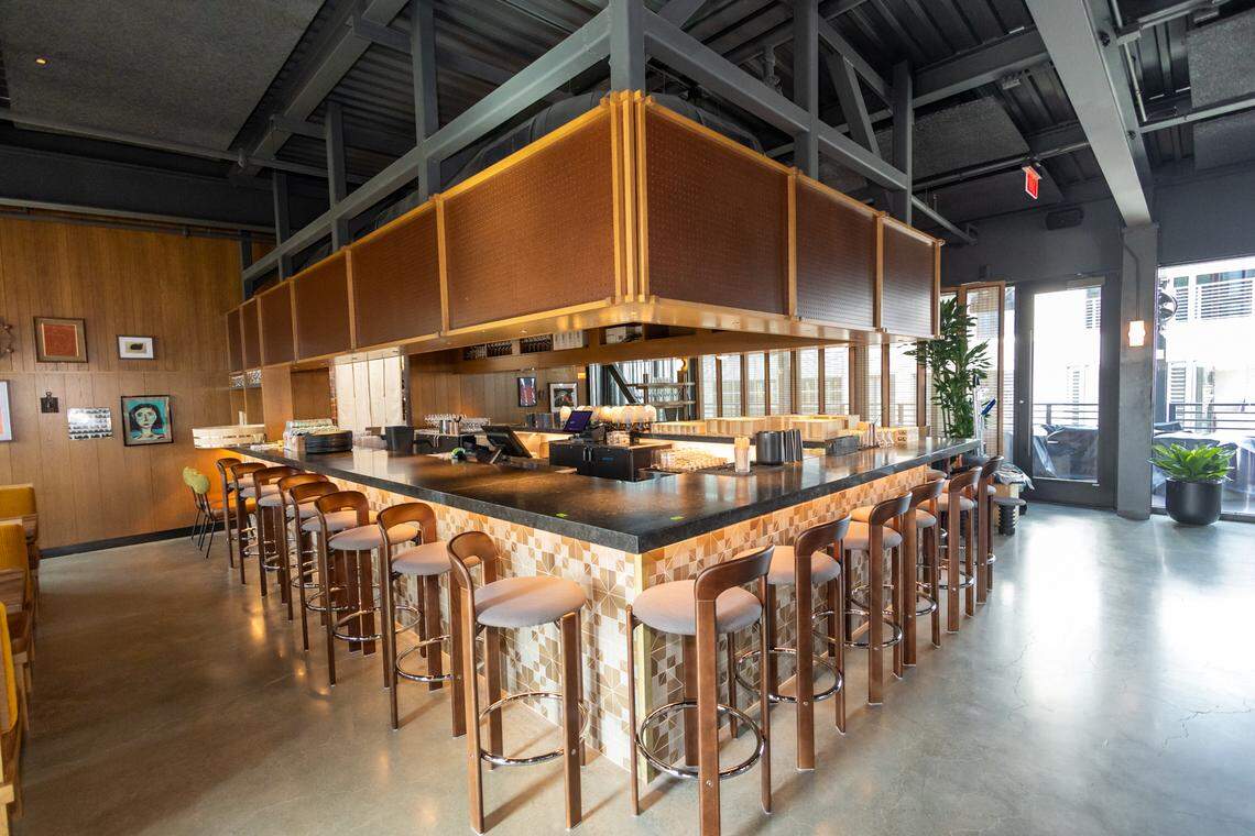 A wide-angle, eye-level indoor shot captures a modern restaurant and bar area with an industrial aesthetic. A large, L-shaped bar with a dark, polished countertop dominates the center. The front of the bar is covered in cream-colored tiles featuring a repeating geometric pattern of light brown triangles. Numerous high-top wooden bar stools with light grey cushioned seats and curved backrests line the perimeter.