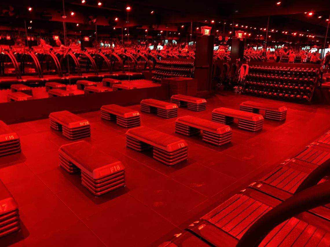 The workout room at Barry’s has treadmills along one wall and strength stations in the center. The dark room with a red light is a signature of the boutique fitness studio.