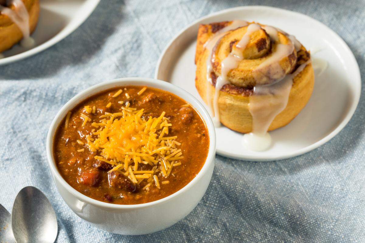 A high-angle shot of a bowl of chili and a cinnamon roll on a light blue tablecloth. The chili, in a white bowl, is topped with a generous pile of shredded cheddar cheese. The cinnamon roll, on a small white plate, is glazed with a thick, white icing that drips down the sides. A spoon is partially visible next to the chili bowl.
