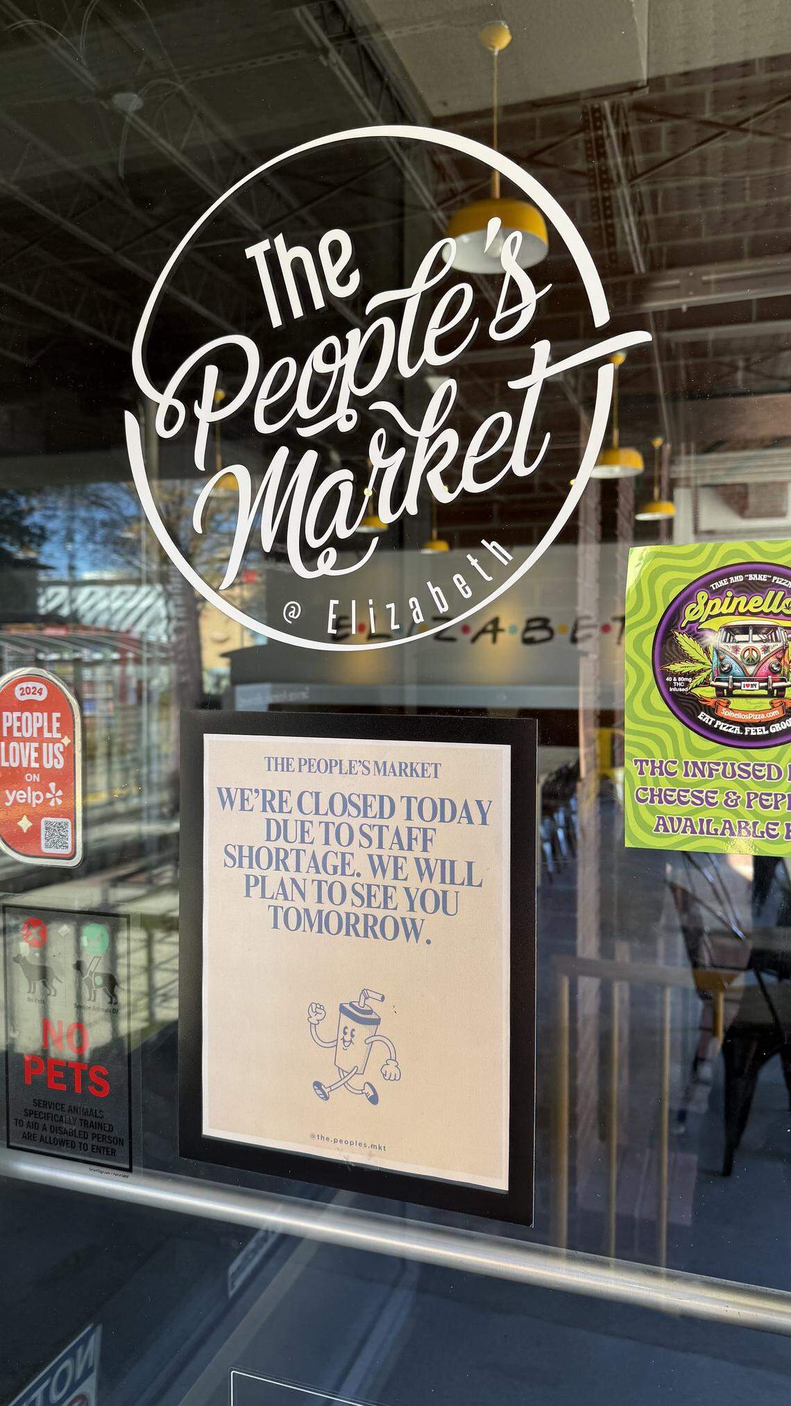 A close-up shot of the glass entrance door to “The People’s Market @ Elizabeth,” which has its name in a white circular logo. A sign taped to the door, just below the logo, reads: “WE’RE CLOSED TODAY DUE TO STAFF SHORTAGE. WE WILL PLAN TO SEE YOU TOMORROW.”