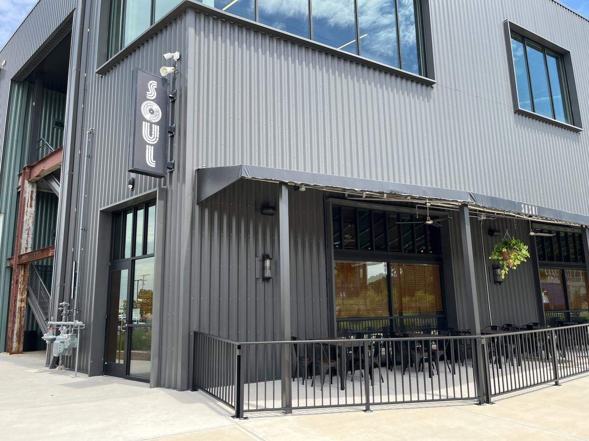 The exterior of Soul Gastrolounge, a restaurant with gray corrugated metal siding. A black vertical sign reads “SOUL” in white lettering. An outdoor patio with black tables and chairs sits under an awning.