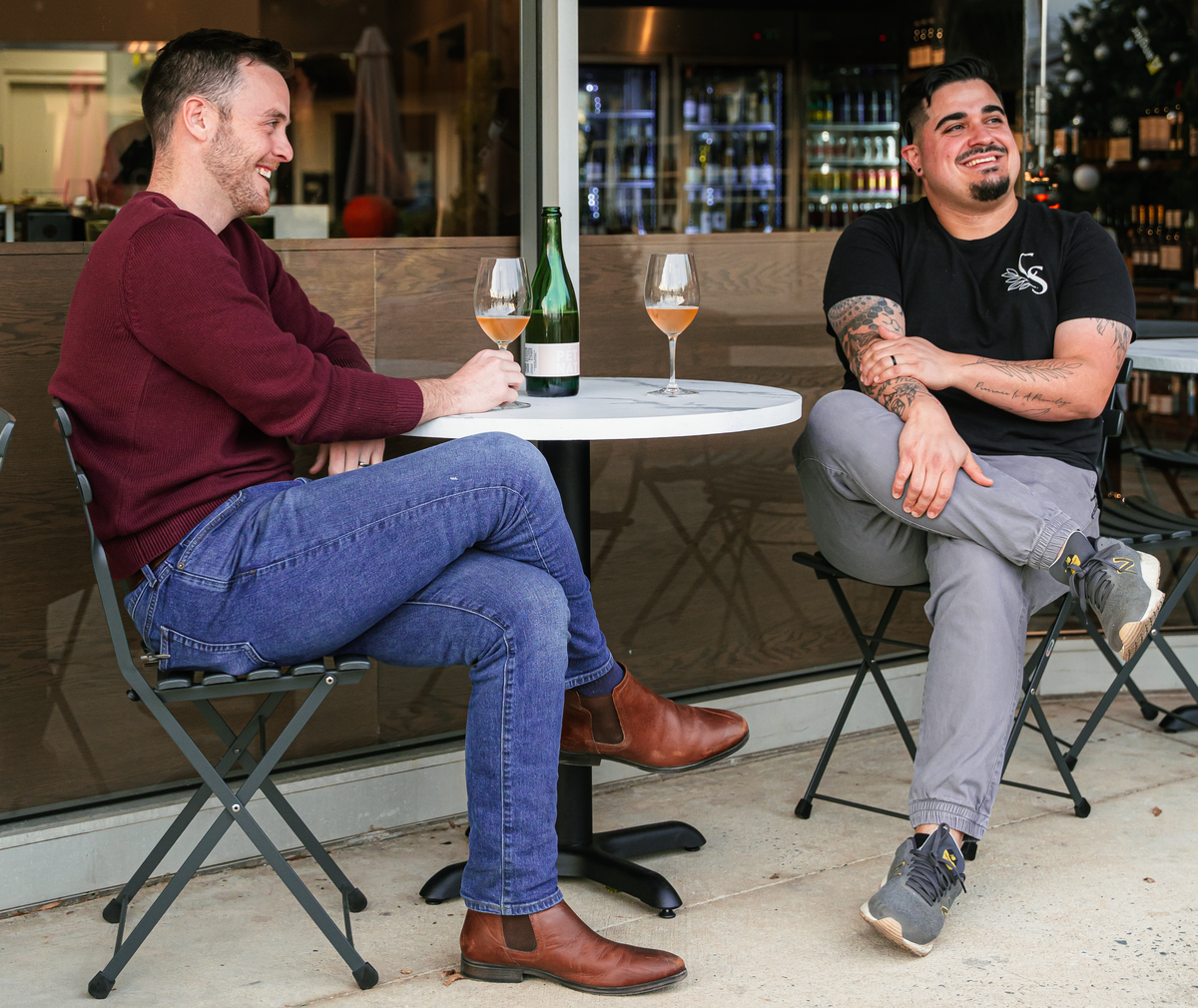 Two people are seated at a small, round white marble table on a patio, laughing and talking. Between them on the table is a green wine bottle and two glasses of rosé or orange wine. The person on the left wears a maroon sweater, blue jeans, and brown Chelsea boots. The person on the right, who has visible arm tattoos, wears a black t-shirt and grey joggers. They are sitting on black folding bistro chairs in front of a glass storefront.