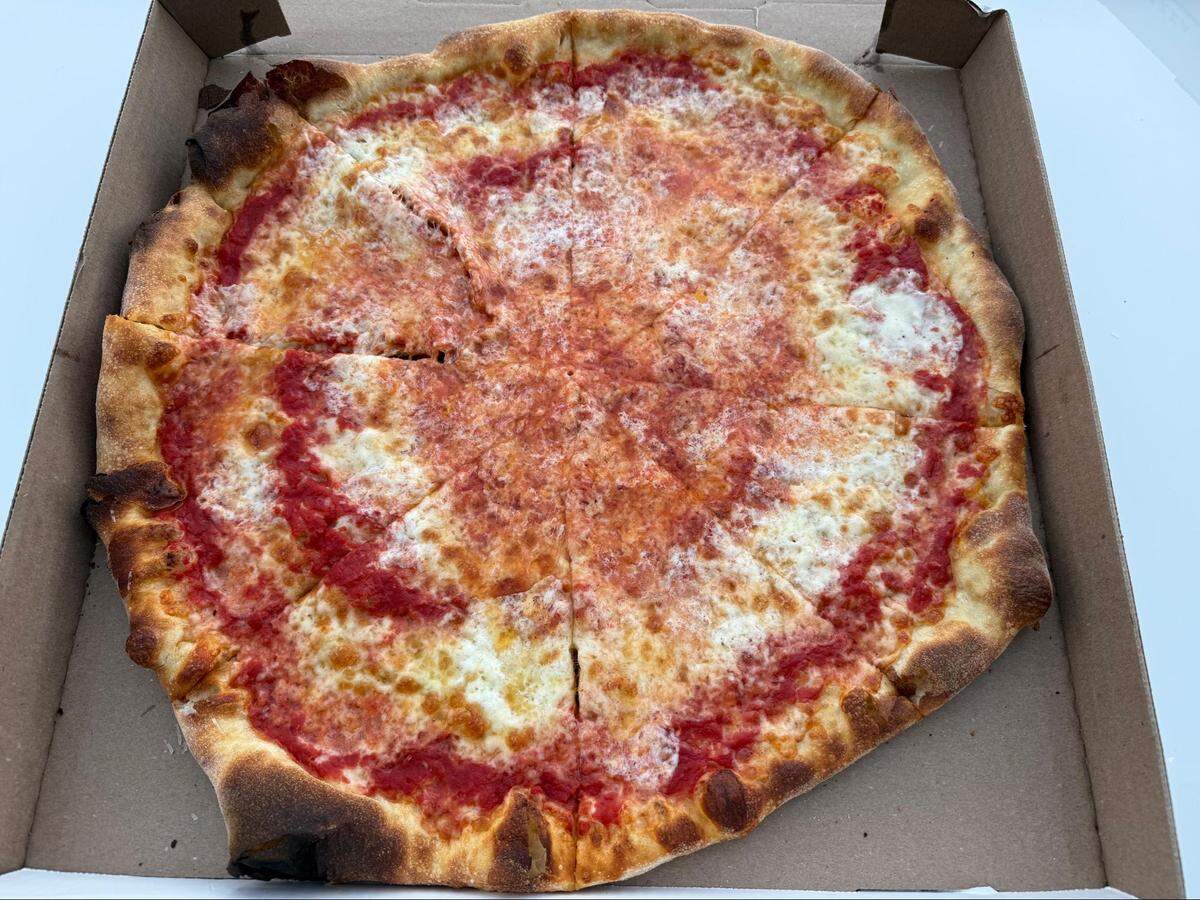 An overhead, close-up shot captures a large, round cheese pizza inside an open cardboard box. The pizza is topped with red tomato sauce and dollops of melted mozzarella, and features a browned, bubbled crust.