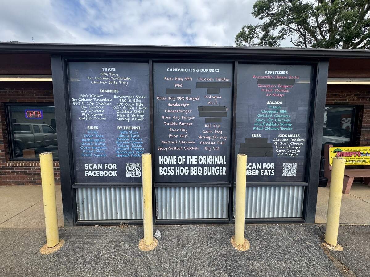 A large outdoor menu displayed across three glass window panes on the side of a brick building. The menu lists various items under categories like “Trays,” “Dinners,” “Sides,” “Sandwiches & Burgers,” “Appetizers,” and “Kids Meals,” with the prominent tagline “Home of the Original Boss Hog BBQ Burger.” Four yellow security bollards stand in the foreground on the asphalt walkway.