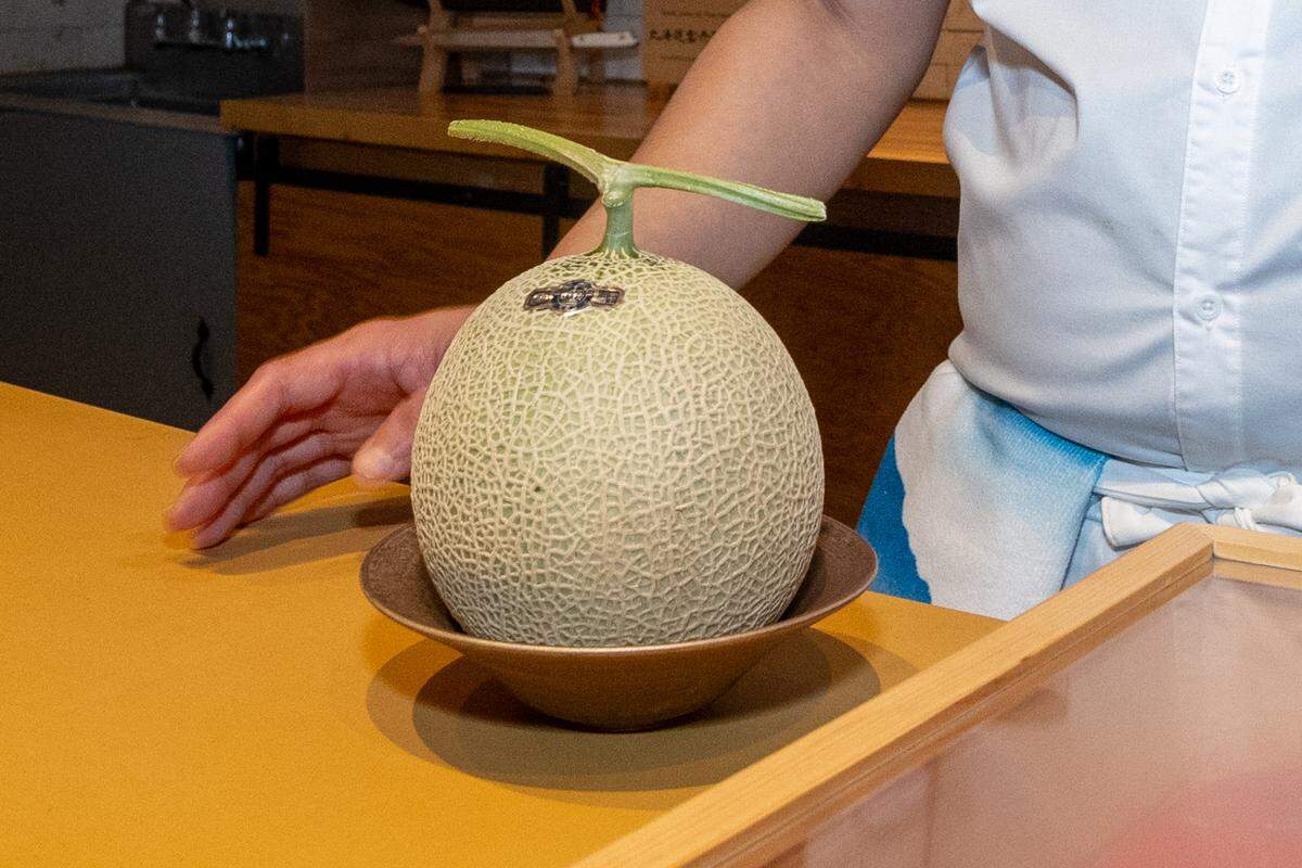 A close-up shot of a whole, premium King Musk Melon with its distinctive netted green skin and T-shaped stem, resting in a shallow bronze bowl on a wooden counter. A chef’s hand and white uniform are partially visible in the background, emphasizing the fruit’s presentation as a high-end ingredient.