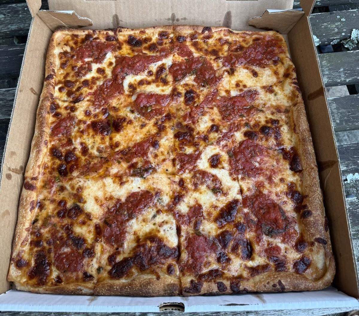 A high-angle shot of a square-cut Sicilian-style pizza in a cardboard takeout box. The pizza has a thick crust and is topped with a generous amount of melted cheese and splotches of red tomato sauce. The cheese is golden-brown and bubbly, with some darker caramelized areas. The pizza is cut into eight rectangular slices. The takeout box is sitting on a dark, rustic wooden surface.