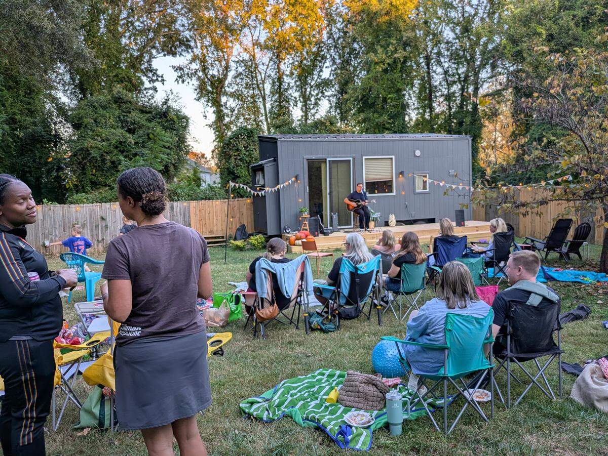A wide shot of an intimate backyard concert at dusk. A man plays an acoustic guitar on a small deck in front of a gray tiny house, while an audience of adults and children watches from lawn chairs and blankets on the grass. In the left foreground, two women stand and talk, facing away from the performer.
