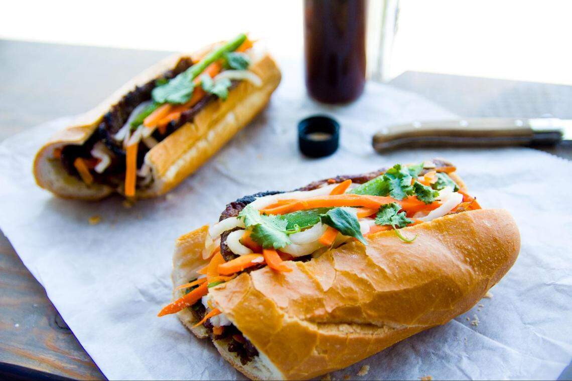 A shallow depth-of-field shot features two banh mi sandwiches resting on a crumpled sheet of white parchment paper on a light wooden surface. In the foreground, one sandwich is in sharp focus, showcasing its crusty baguette filled with grilled meat, pickled carrots, white onions, and fresh cilantro. Another similar sandwich is visible, blurred, in the background. A dark glass bottle and a knife with a wooden handle are also out of focus in the upper right background.