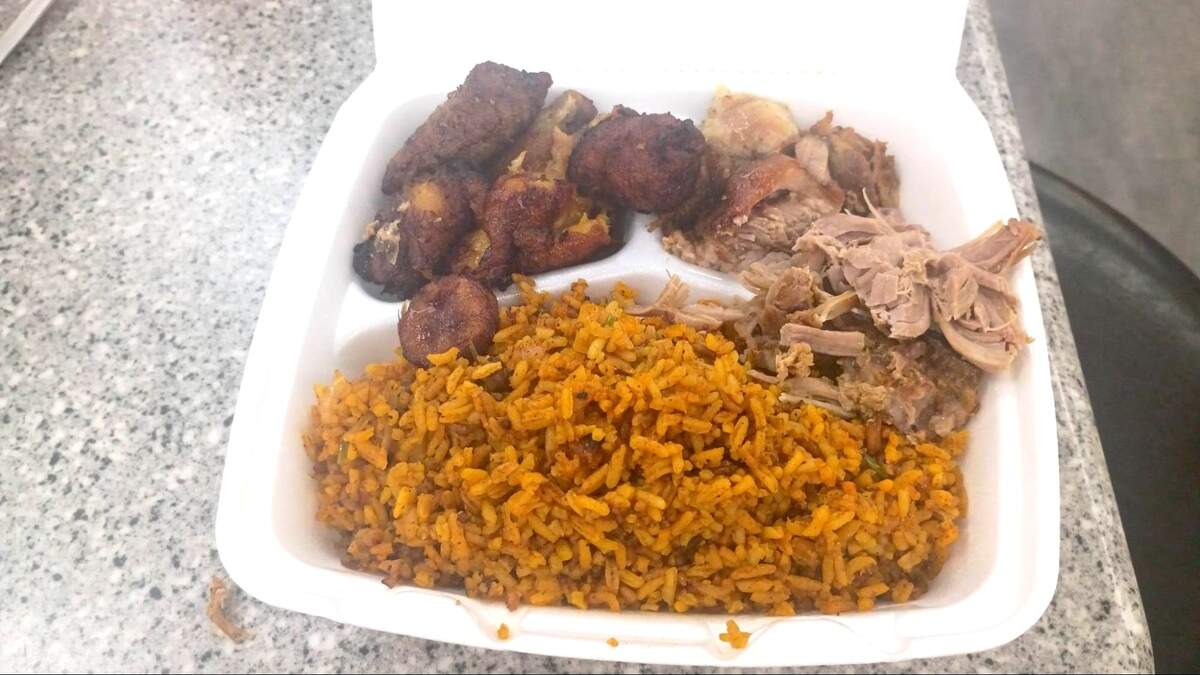 A white styrofoam takeout container filled with a large portion of yellow seasoned rice (arroz con gandules), shredded slow-roasted pork (pernil), and several pieces of fried sweet plantains (maduros).