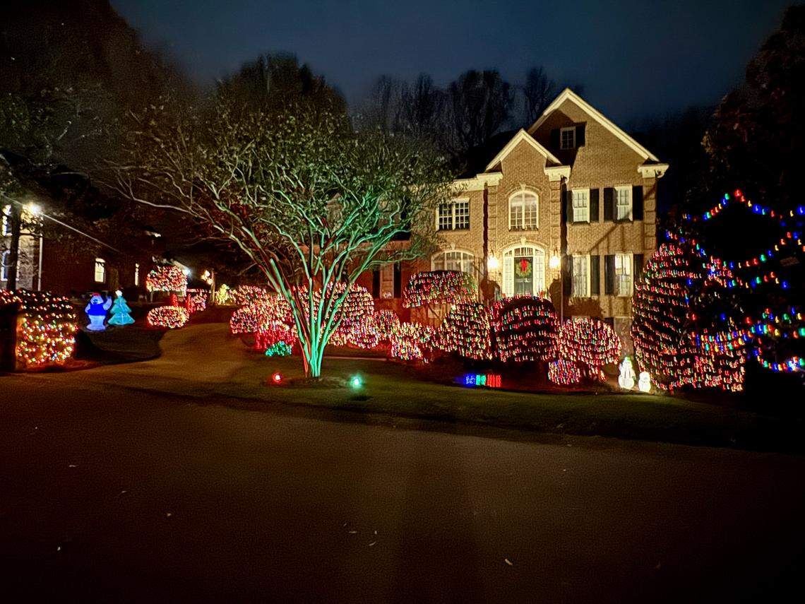 A nighttime photograph showing a large, traditional brick house brightly lit up for the holidays. The front yard is densely covered in colorful lights, including red lights on the bushes, multicolored string lights on the trees, and a central tree illuminated with a vibrant green light. Small illuminated snowmen figures are also visible near the sidewalk.