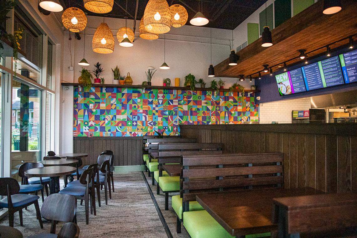 A bright and colorful photograph of the interior of a modern restaurant. The dining area features a row of wooden booths with bright green cushions and several round tables with dark chairs. A vibrant, geometric mural in various colors covers a wall above the booths. Hanging lights with woven shades and black metal fixtures illuminate the space. The counter and a digital menu board are visible in the background, and large windows on the left provide natural light.