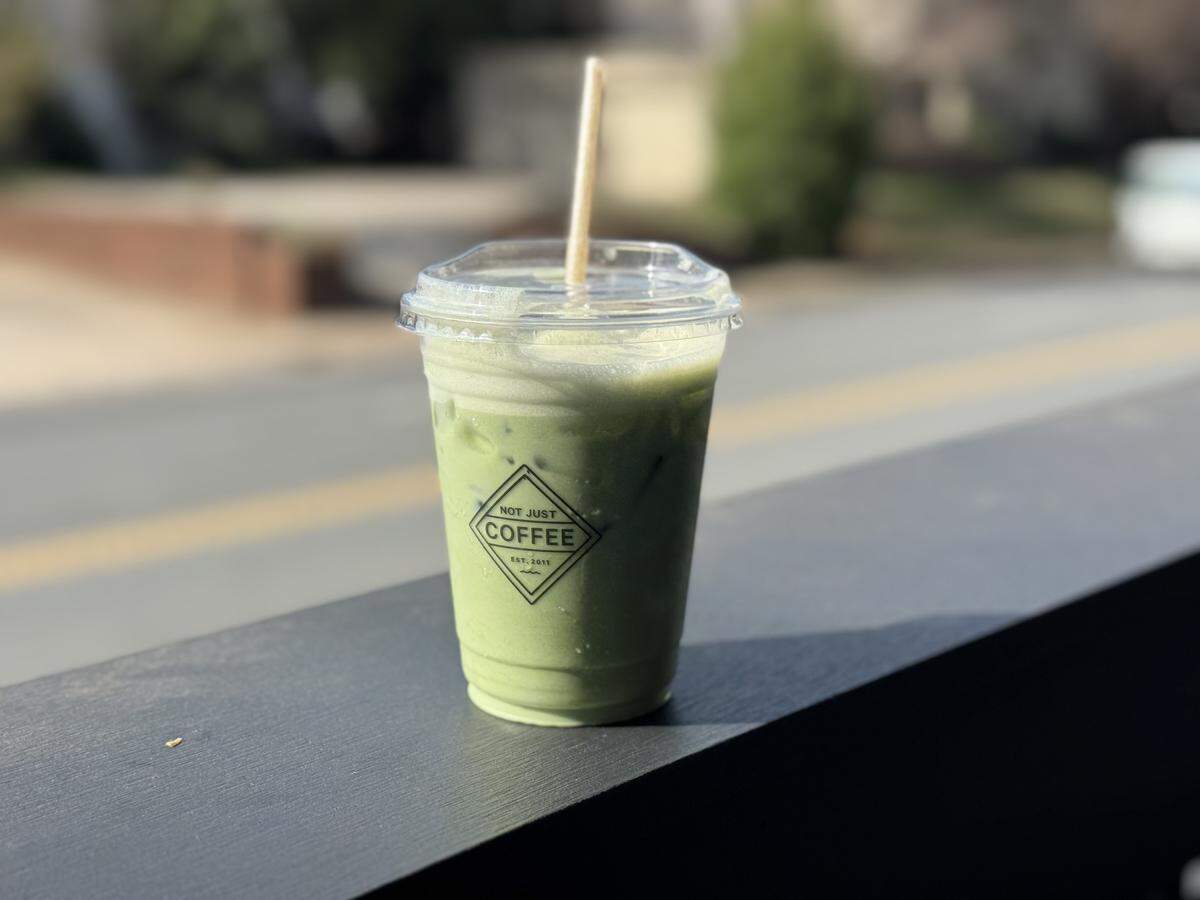 An iced matcha green tea latte in a clear plastic cup with a lid and a straw. The drink is a vibrant pastel green with a frothy top, sitting on a dark ledge with a blurred street and trees in the background.