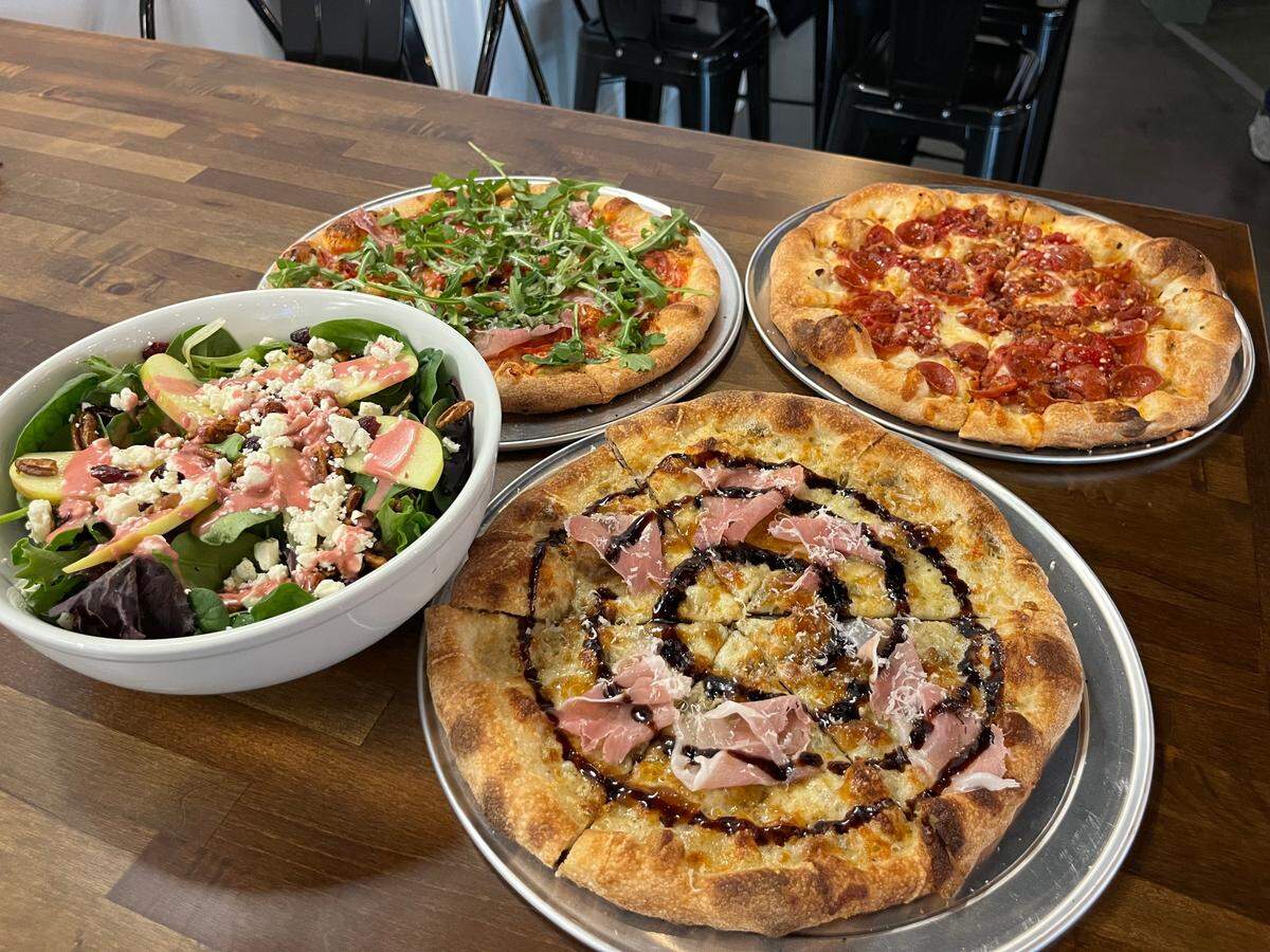 A high-angle view of a dining table featuring three artisanal pizzas and a large salad. The salad in the foreground is a mix of leafy greens topped with sliced apples, pecans, feta cheese, and a vibrant pink dressing. The pizzas include one topped with fresh arugula and prosciutto, a classic pepperoni and tomato pizza, and a white pizza drizzled with a balsamic glaze swirl and topped with thin ribbons of prosciutto.