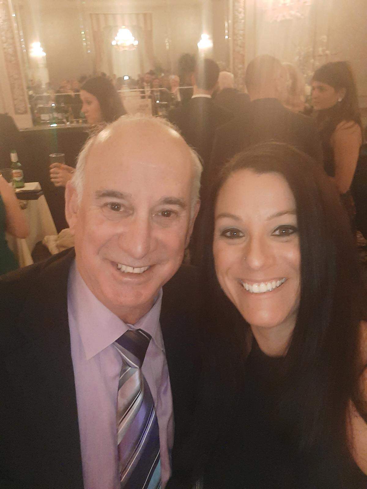 A candid selfie showing an older man and a younger woman smiling broadly at a formal gathering or banquet. The man is wearing a suit, lavender shirt, and a striped tie, and the woman has long dark hair and is wearing a black top. The background is a brightly lit, ornate room with many other guests in attendance.