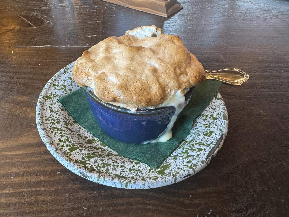 A blue ceramic ramekin filled with a dessert topped with a high, golden-brown toasted meringue, served on a green speckled plate over a rustic wooden table.