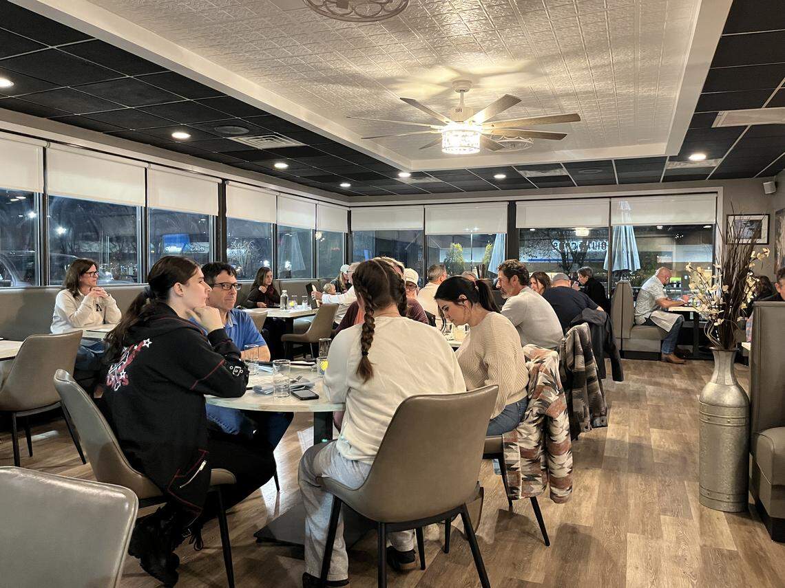 An eye-level, wide shot capturing the interior of a moderately busy restaurant dining room in the evening. Several groups of patrons are seated at various tables and booths, engaged in conversation. The room features a wood-grain floor, gray upholstered chairs and booths, large windows, and a dark ceiling with white decorative tiles.