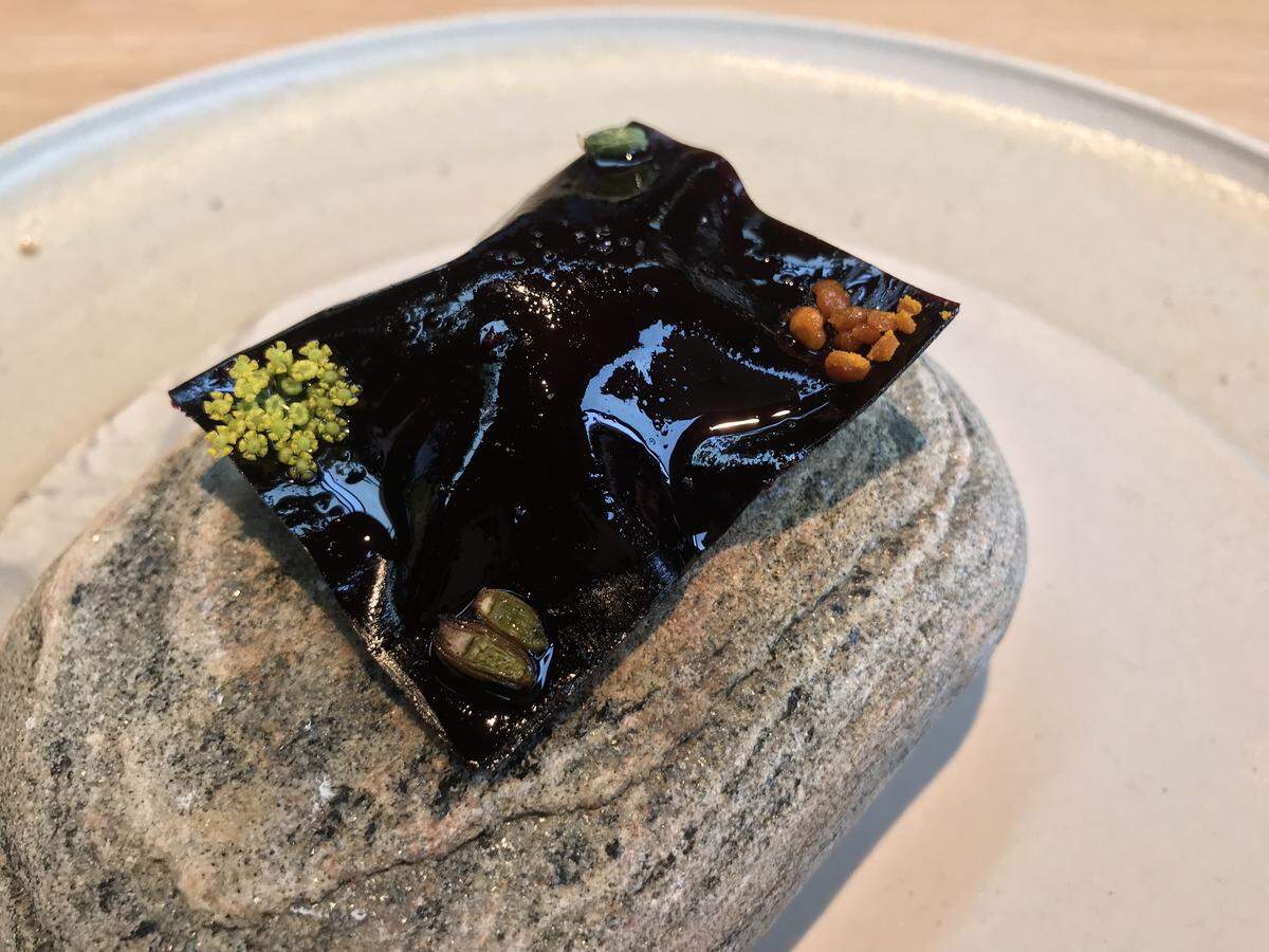 A thin, glossy, dark purple fruit leather square draped over a smooth grey river stone. The leather is delicately garnished with tiny yellow flowers and small orange seeds.