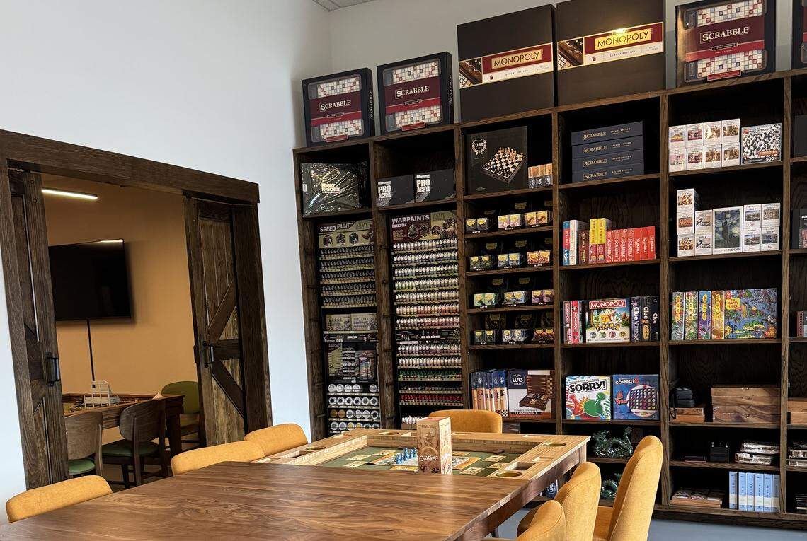 A warm, inviting game room featuring a large, dark wooden shelf unit against a white wall, filled with board games, miniature wargaming supplies, and paint sets. Prominent items include upscale versions of games like Monopoly and Scrabble on the top shelf, various paint bottles organized vertically, and classic board games like Sorry and Trivial Pursuit below. In the foreground is a wooden gaming table with a sunken playing area (possibly a board game or puzzle table), surrounded by orange upholstered chairs. To the left, a wooden barn-style sliding door opens to another room with more tables and a wall-mounted TV.