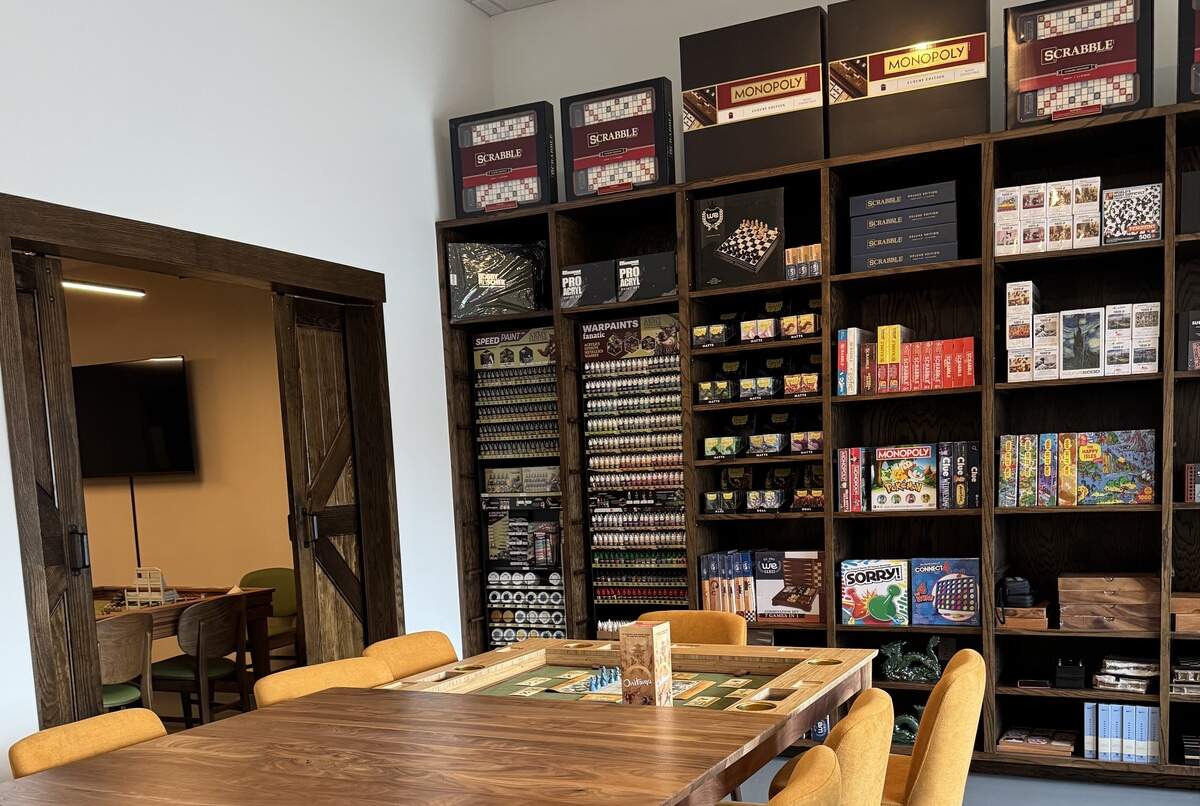 A warm, inviting game room featuring a large, dark wooden shelf unit against a white wall, filled with board games, miniature wargaming supplies, and paint sets. Prominent items include upscale versions of games like Monopoly and Scrabble on the top shelf, various paint bottles organized vertically, and classic board games like Sorry and Trivial Pursuit below. In the foreground is a wooden gaming table with a sunken playing area (possibly a board game or puzzle table), surrounded by orange upholstered chairs. To the left, a wooden barn-style sliding door opens to another room with more tables and a wall-mounted TV.