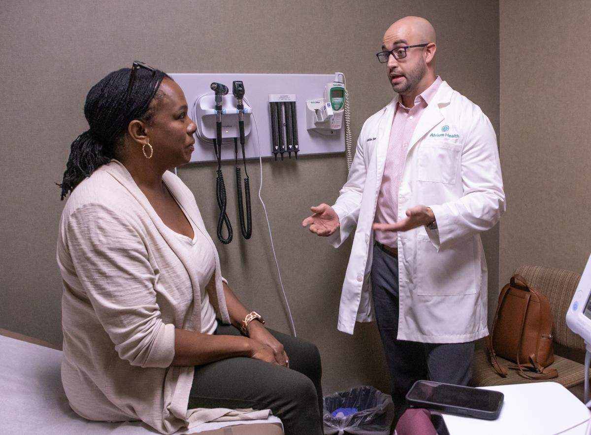 A smartphone using new AI tech sits beside Atrium Health Primary Care Cabarrus Family Medicine Dr. Austin Bush as he meets with patient Valerie Burton. The tech documents the conversation, which is then summarized and loaded into the patient’s electronic medical record.