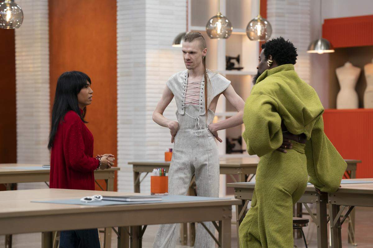 Three people are standing in a circle looking at each other. Two have their hands on their hips. One is wearing red, one grey and one lime green. Beige folding tables surround. A pair of sunglasses sits in the corner of one next to a notebook. The room has a bright orange wall with white tiles. Three lights hang down from above and a couple of mannequin busts are in a cutout on the wall.