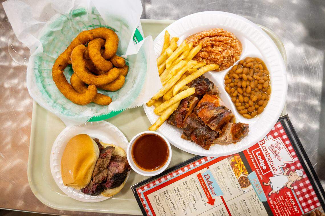 A top-down view of a meal from Red Bridges Barbecue Lodge. A large plate is filled with chopped pork barbecue, french fries, baked beans, and red slaw. This is accompanied by a separate basket of onion rings, a brisket sandwich, a cup of sauce, and the restaurant’s paper menu.