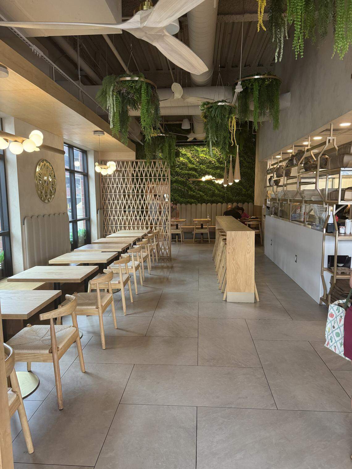 A photo of a modern and stylish cafe interior. There are several long tables with wooden chairs arranged for seating. The decor includes light wood tones, hanging plants, a large green moss wall, and modern light fixtures.