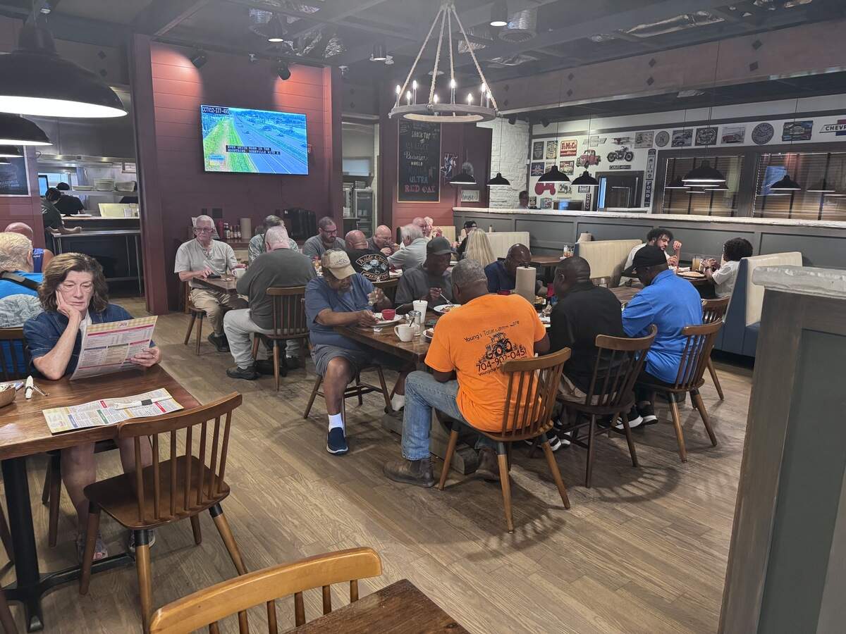 The busy dining room of a casual restaurant filled with patrons seated at wooden tables and booths, enjoying meals. Warm lighting, rustic wooden accents, a hanging wheel-style chandelier, and a TV mounted on a reddish-brown wall create a cozy atmosphere.