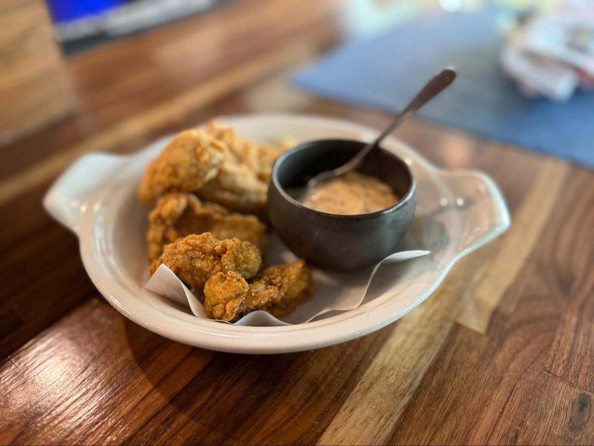 A plate of golden-brown, crispy fried oysters resting on white parchment paper. A small black stoneware bowl sits on the side containing a creamy, speckled dipping sauce and a small silver spoon.