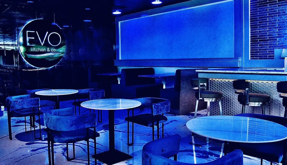 The modern interior of a restaurant dining area is bathed in dramatic, vibrant blue neon lighting. Round tables with white tops are surrounded by plush, velvet-textured blue chairs, while dark booth seating lines the back wall. On the far left, a circular glowing sign reads “EVO kitchen & co.” against a dark, textured wall surface.