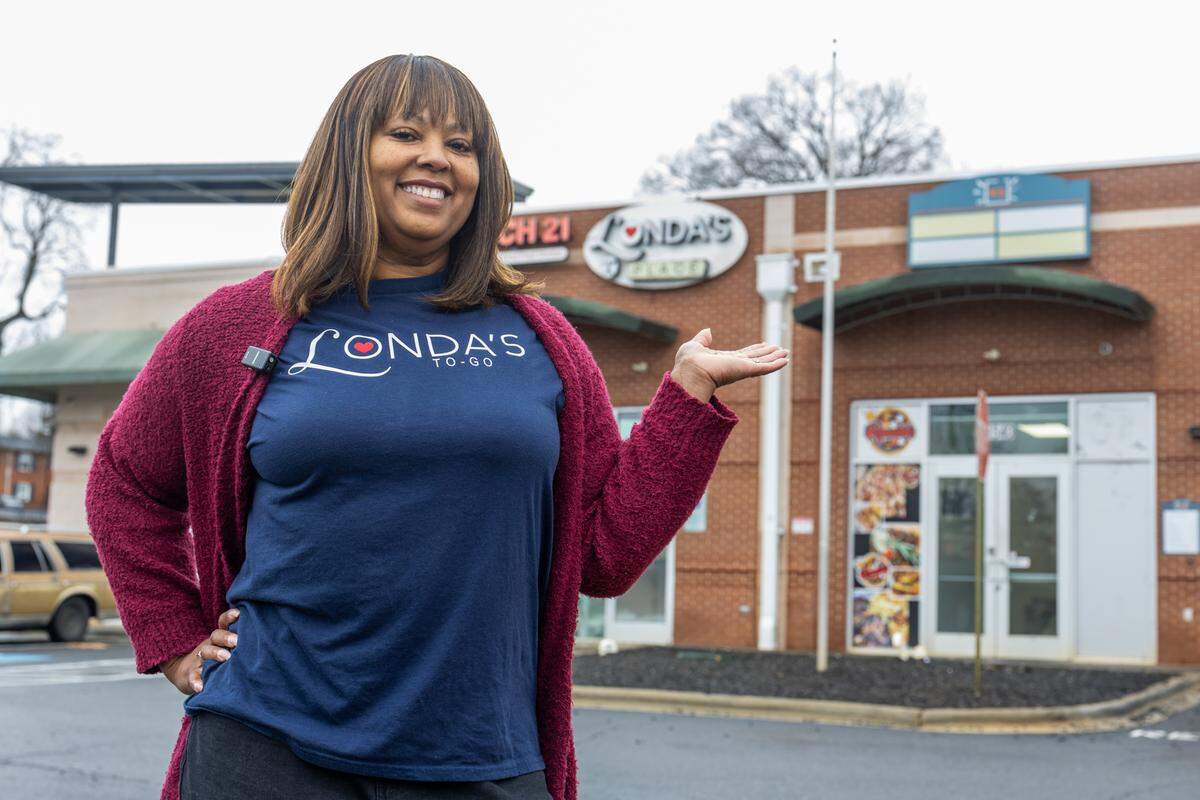 A woman with brown hair and a bright smile stands in a parking lot, wearing a burgundy cardigan and a navy blue t-shirt that reads ‘Londa’s To-Go’ with a red heart over the ‘o.’ She is gesturing with her open hand toward a brick storefront in the background, which has a partially visible circular sign for ‘Londa’s Place’ or ‘Londa’s.’