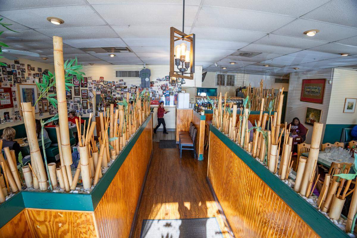 A high-angle, wide shot looking down the main walkway of the Lang Van restaurant. The aisle is flanked by low, wood-paneled dividers, which are filled with thick, decorative bamboo stalks, separating the walkway from the dining booths on either side. The back wall is famously covered in a dense collage of framed photos and memorabilia. Patrons are seated in the dining areas, and a staff member is visible in the background.