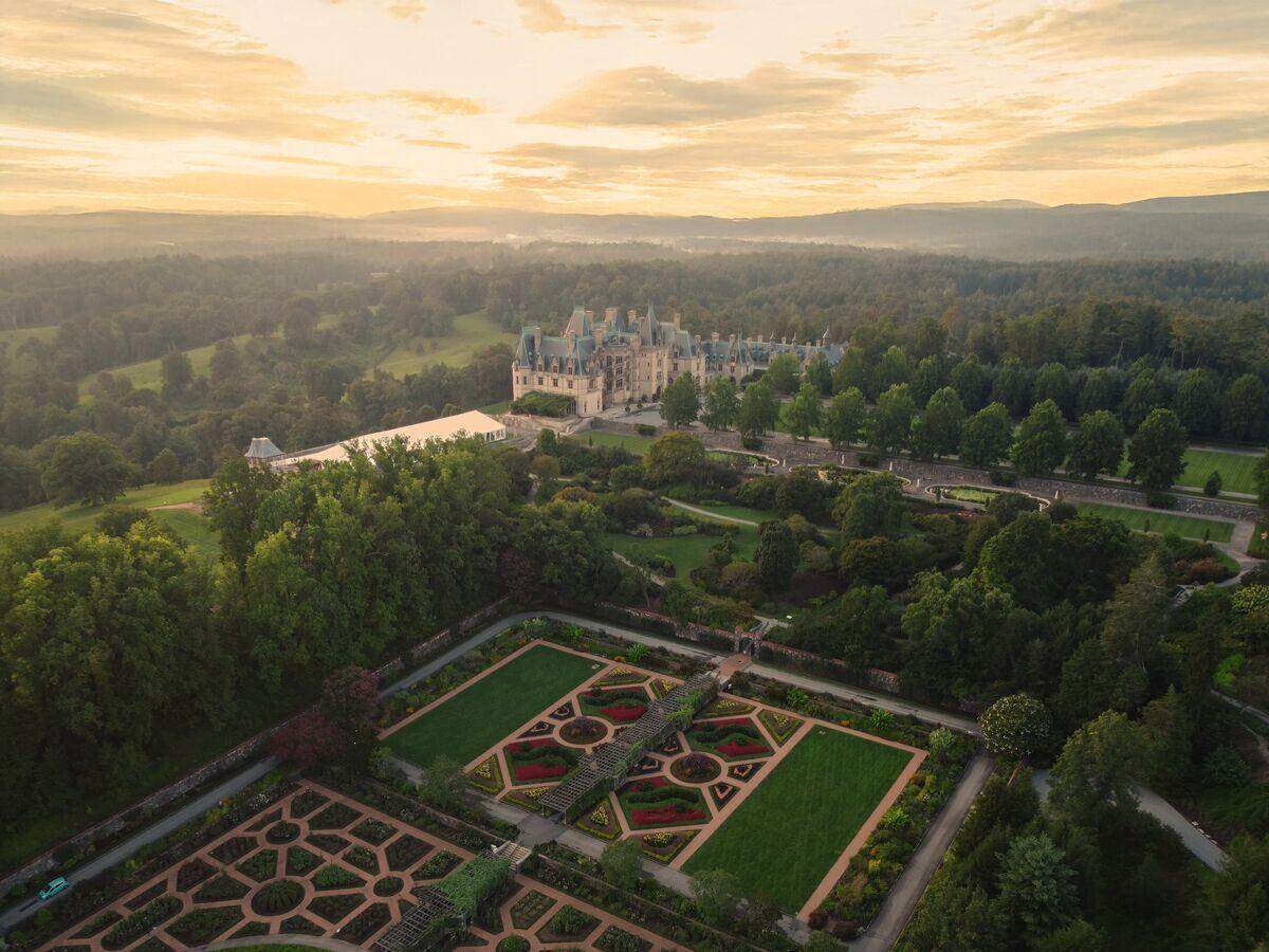 A sweeping, golden-hour aerial shot of the Biltmore Estate. The foreground is dominated by the geometric patterns of the formal gardens. Beyond the gardens, the massive, French-style Biltmore House is nestled in a vast forest, with the hazy Appalachian Mountains on the horizon.