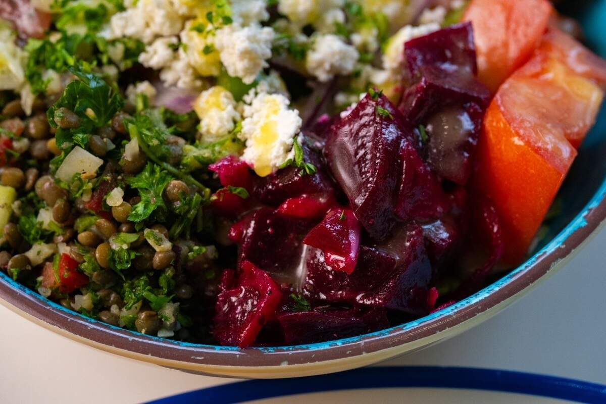 A tight, macro shot focuses on the vibrant ingredients inside a textured turquoise and white bowl. Glistening chunks of deep purple beets sit adjacent to bright red diced tomatoes and a fresh green lentil salad packed with minced herbs. The dish is finished with a generous topping of crumbled white feta cheese drizzled with olive oil.